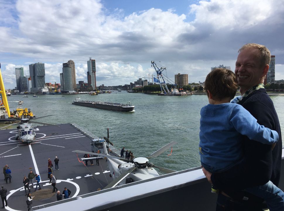 father and son standing on the deck of a ship with helicopters and boats passing by in rotterdam