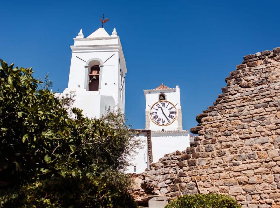 white church towers with bel and clock behind a remaining castle wall in tavira portugal