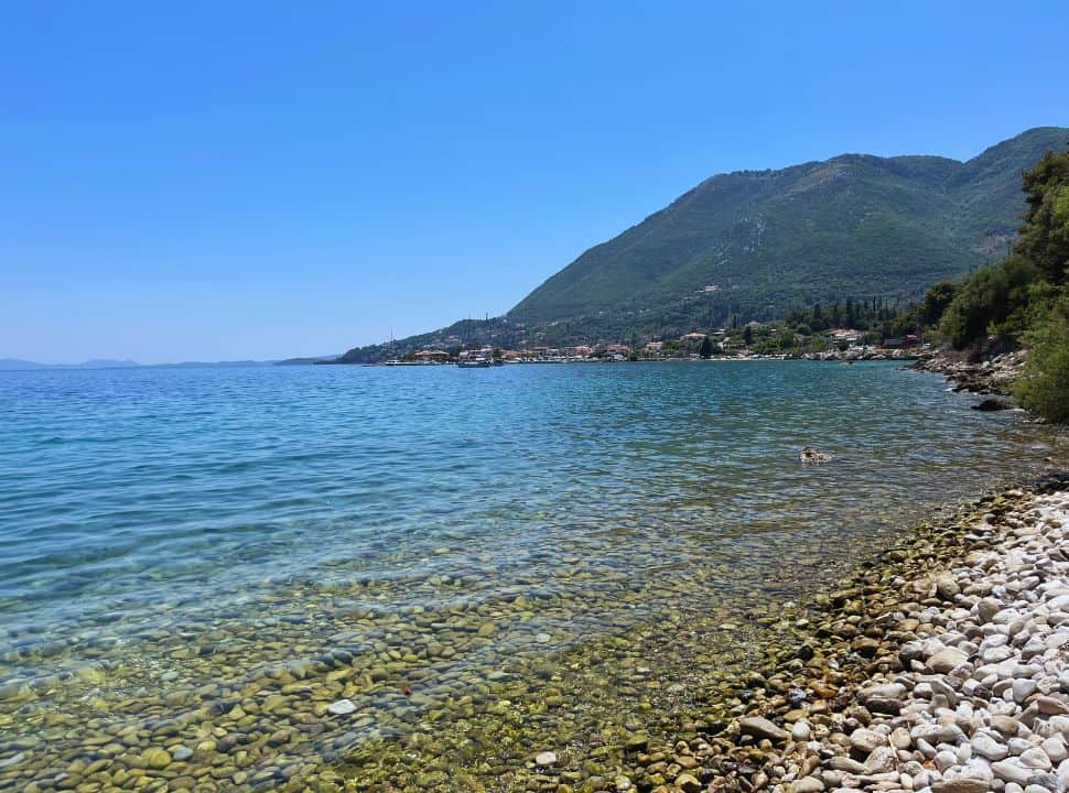 large pebbled beach with clear water and in the back ground hills and a coastal village