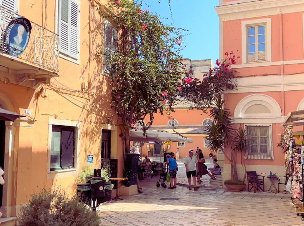 small square surrounded by pink and yellow building, with flowers. 