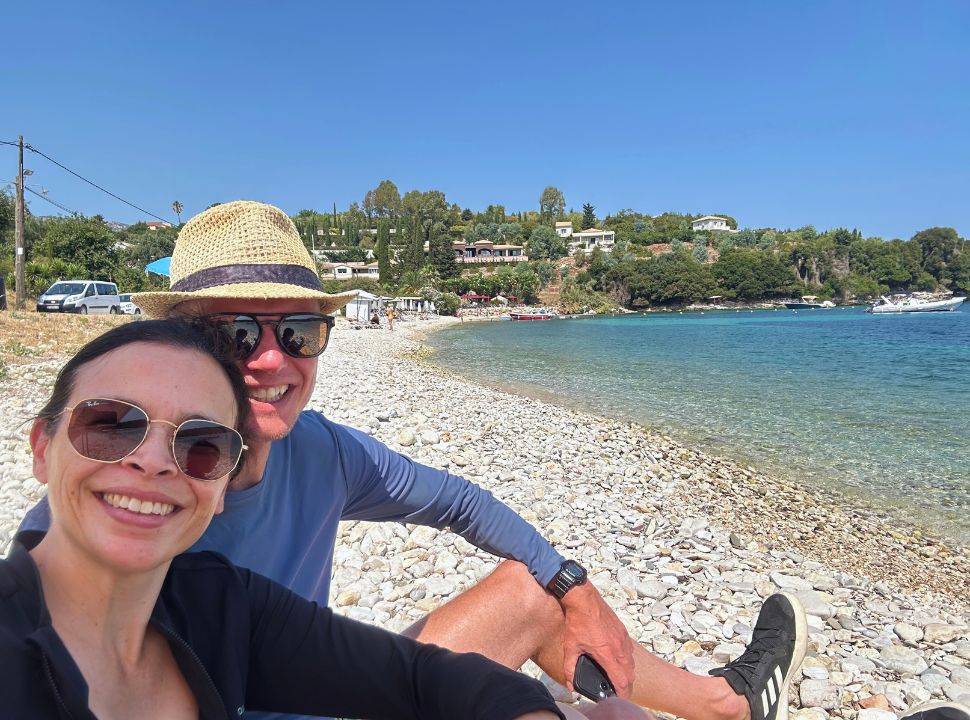 couple posing for a picture on a pebbled beach with accommodation set on the hills on the background