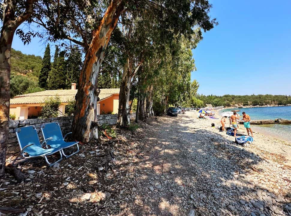 villa located under the trees along a pebbled beach where people are enjoying themselves