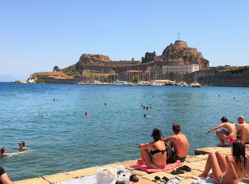 people sitting on the edge of the water and swimming in the water close to the old fortress in corfu town