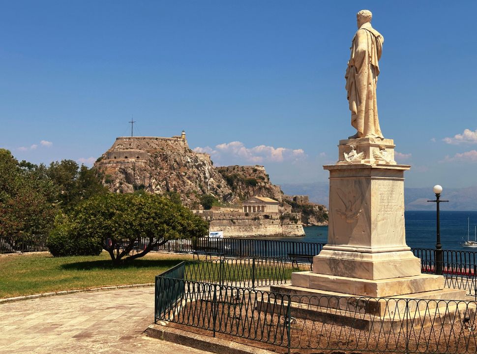 marble statue of a man located in a small park with the old fortress of corfu in the background