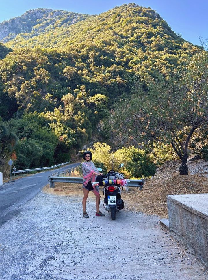 woman standing next to a motorscooter parked next to a mountain road on corfu island