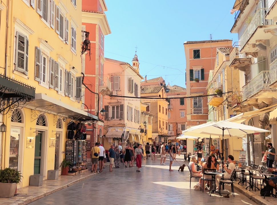 small street in Corfu town with yellow, ochre and light red buildings with a balcony and shutters. In the far distance there is a church tower rising above the rooftops