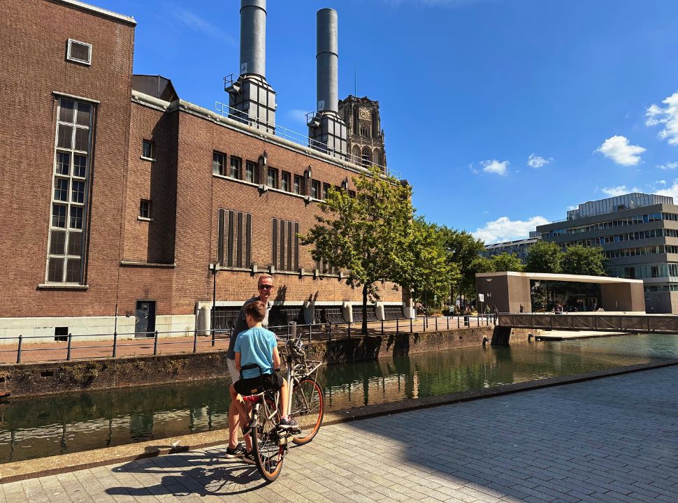 father and son on the bike standing in front of a factory like building with the tip of laurens church in the back