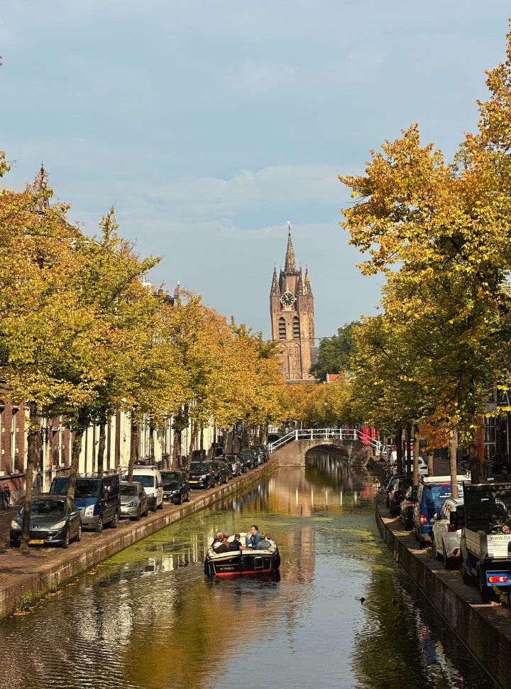 small canal boat navigating through a small canal in Delft with the old church in the background, cars are parked along the canal