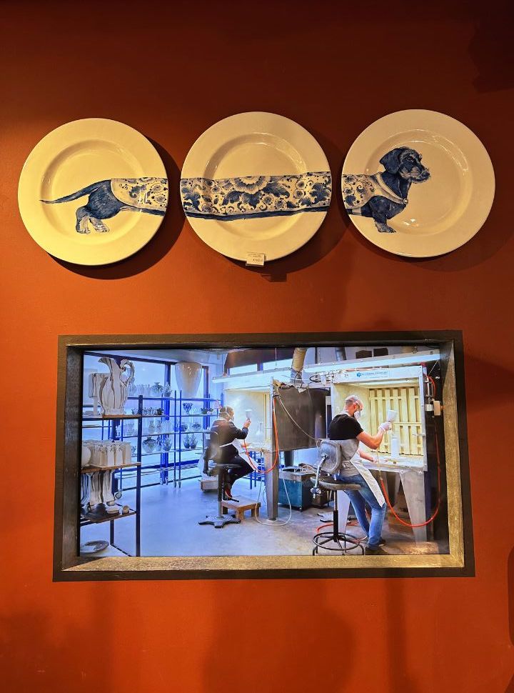 three plates displayed on the wall with a of a dachshund in delft blue with underneath a tv screen where you can see how the ceramics are prepared and painted