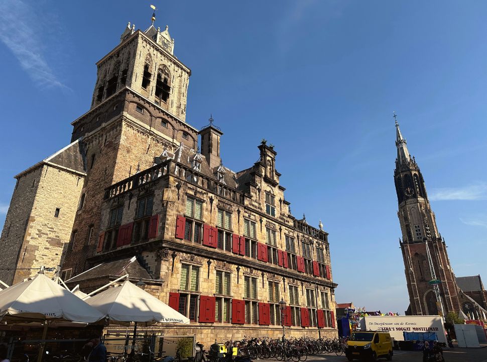 the town hall in delft is a symmetric building with red shutters, a tower in located right in the center of the building. the new church is just across the square