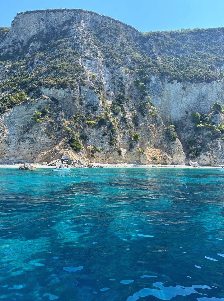 various shades of blue water with white cliffs with vegetation in the back. A small boat is anchored near a deserted beach.