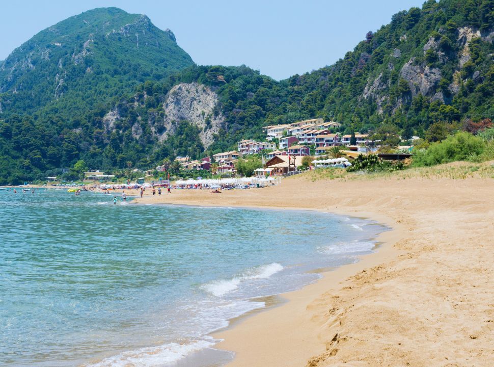 view of the beach with soft sand, calm water and in the far distance a tourist crowd and accommodation. The mountains surrounding the beach is covered with trees. 