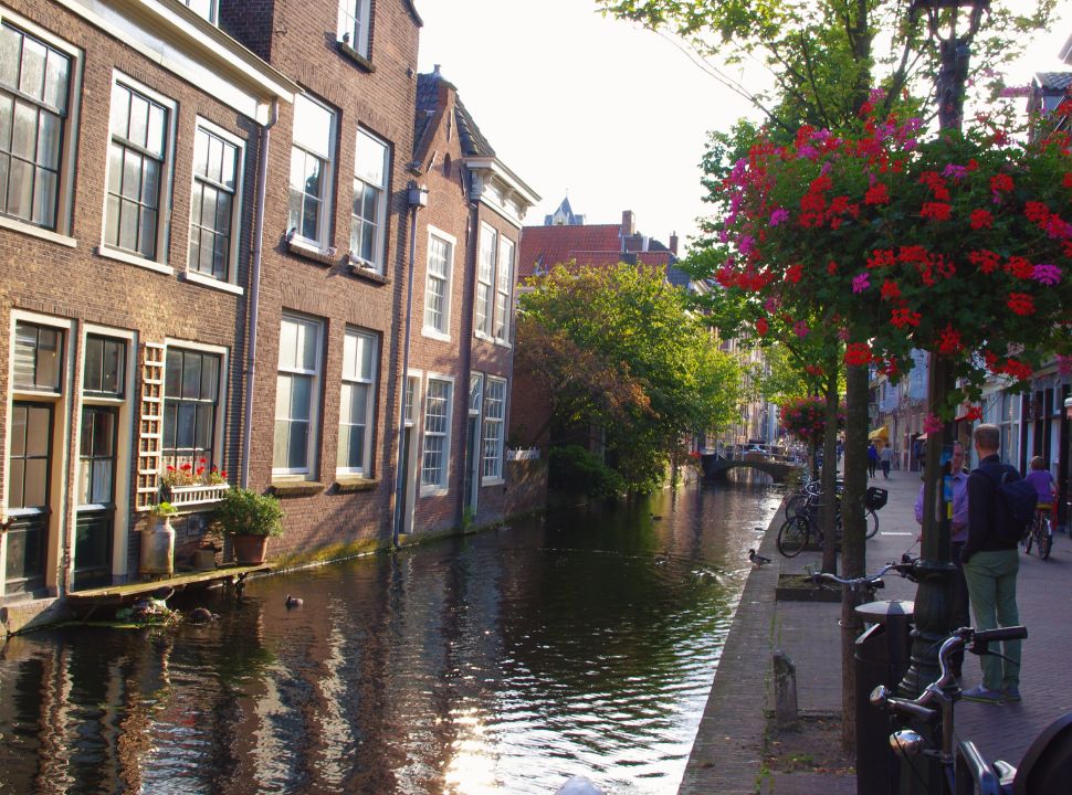 historic houses built at the water of the canal, with a street on the other side of the canal. Flower decorate the streets in Delft