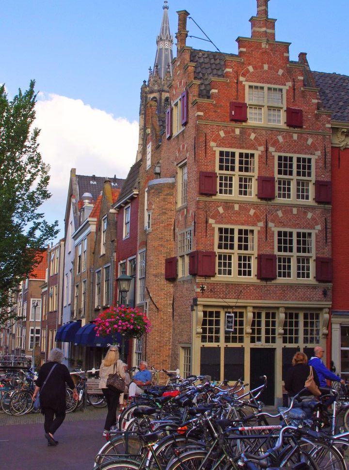 a old historic building with many windows with red shutters located at a canal and a corner of the street, in front are many biked parked