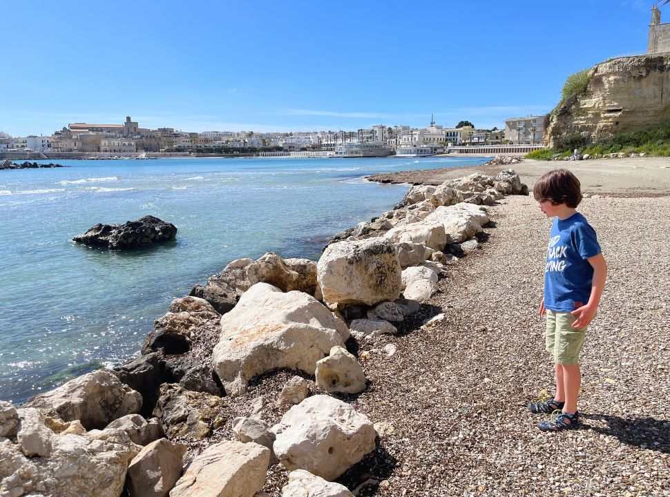 boy watching the waves hit the small rocks at Otranto beach, the city is visible in the back