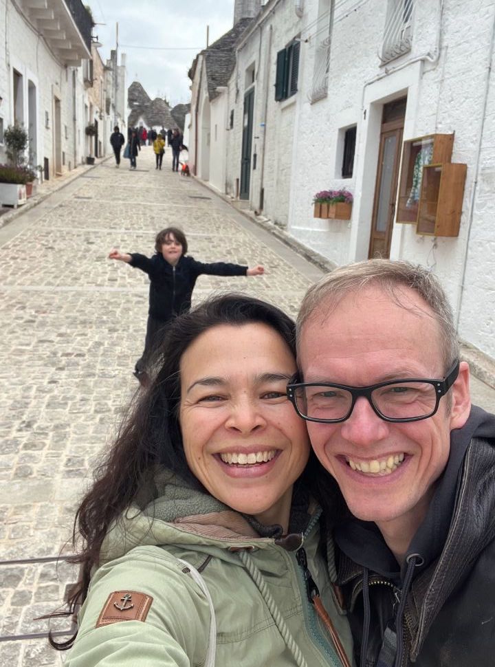 couple posing in a narrow street with trulli houses in Alberobello Puglia, while their son is running towards them in the back