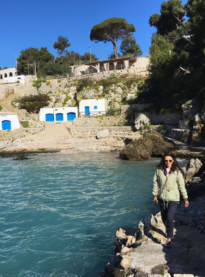 woman walking along the path next to the cliffs and water with in the back sheds for the fishermen and above a restaurant in puglia italy