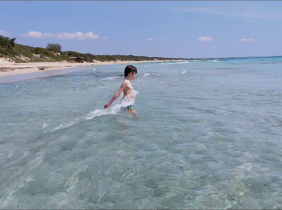 boy wading into the clear ocean with small waves with dunes in the background in Puglia Italy