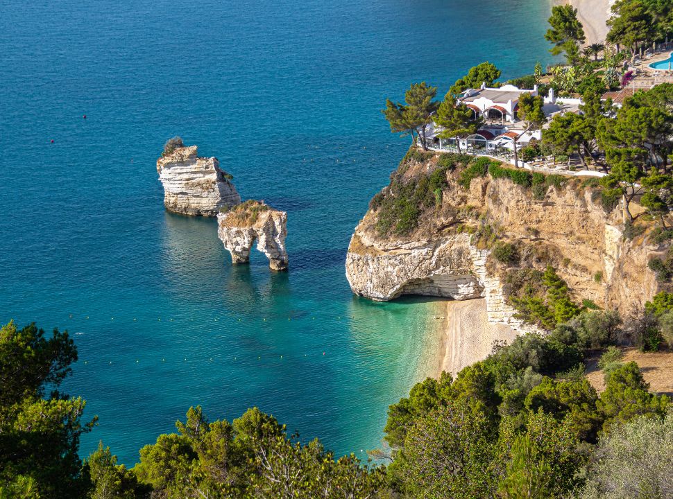 view of cliff beach with tall rock arches in the water, little pebbled beach with blue turquoise water in gargano peninsula Puglia