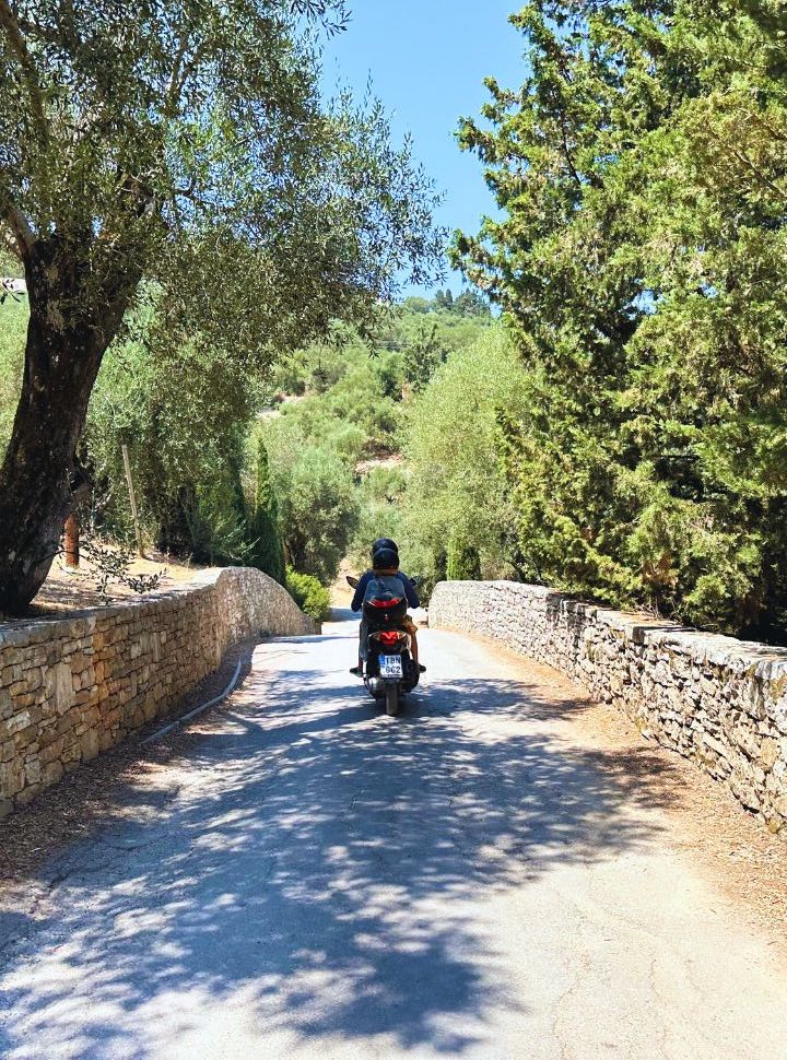 father and son riding a motor scooter along a small street with green vegetation in corfu island