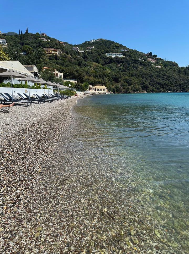 clear water at a pebbled beach with sunbeds with in the back hills covered with trees and dotted with accommodation along the east coast of corfu