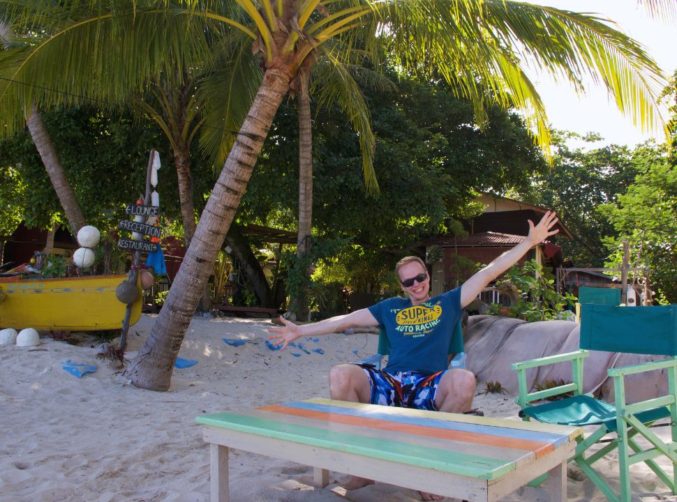 man posing for a picture at a beach restaurant with palm trees and white sand in malaysia