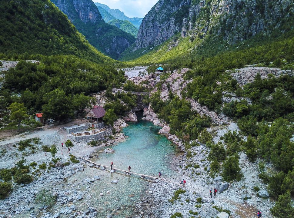 people crossing a small bridge over a crystal clear river flowing through a mountain gorge in Albania