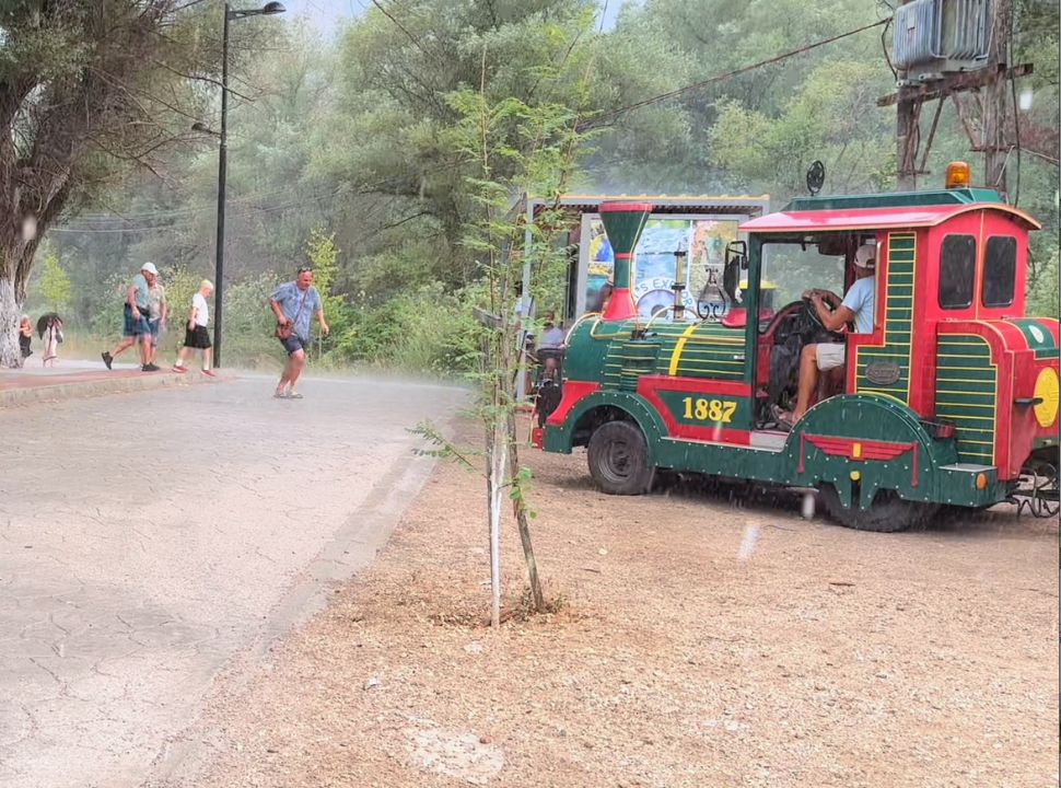 red and green road train parked on the side of the road, the driver is waiting for passengers who are running to the train to avoid the rain at blue eye Albania