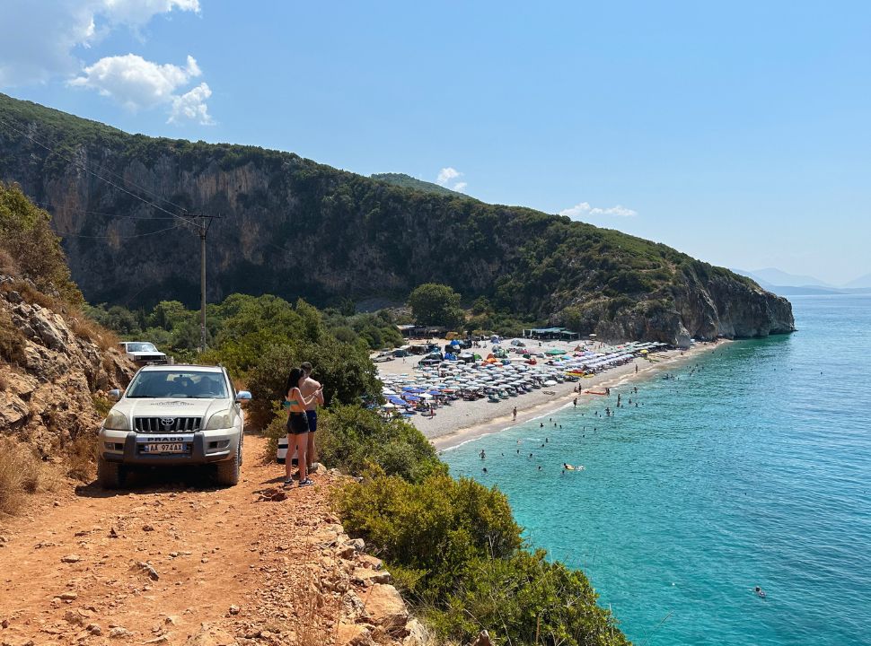 red dirt road leading to a beach cove with beautiful water and sand. A car is passing a couple along this narrow road at Gjipe Beach Albania