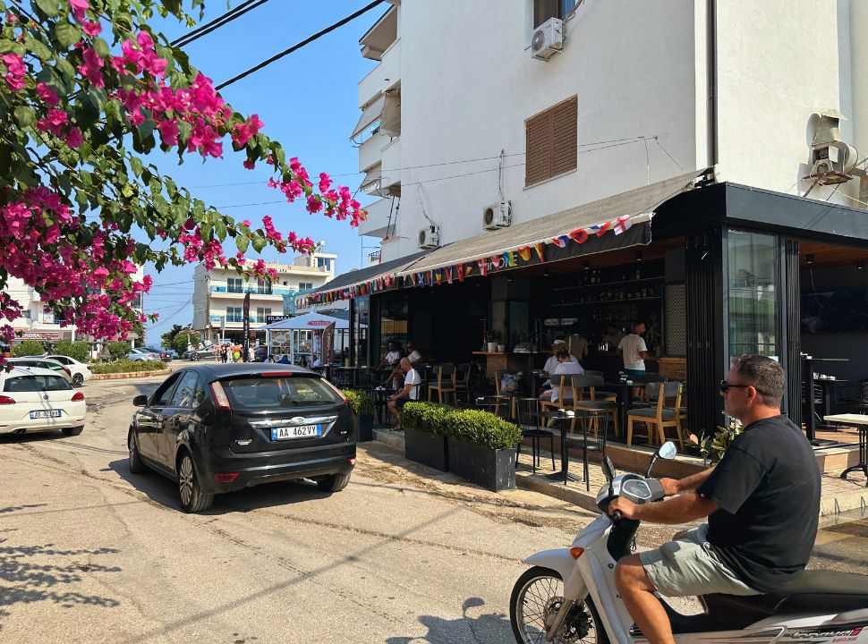 street in ksamil with a restaurant, cars and a motorscooter passing by