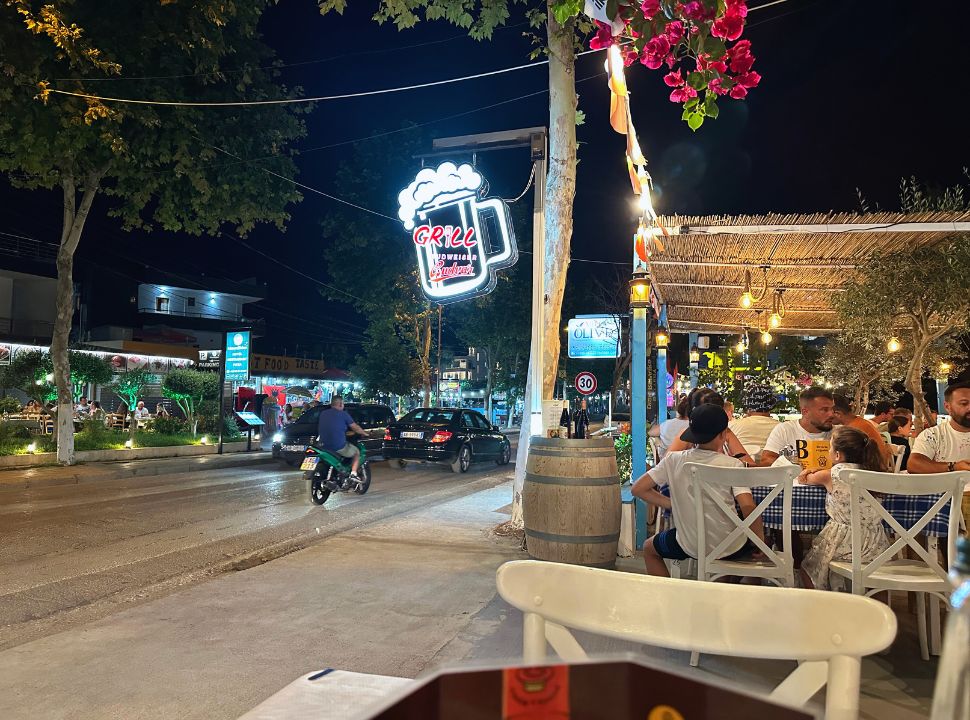 main street of Ksamil by night, cars and a motorbike are passing while on both sides of the street are restaurants where people are having dinner