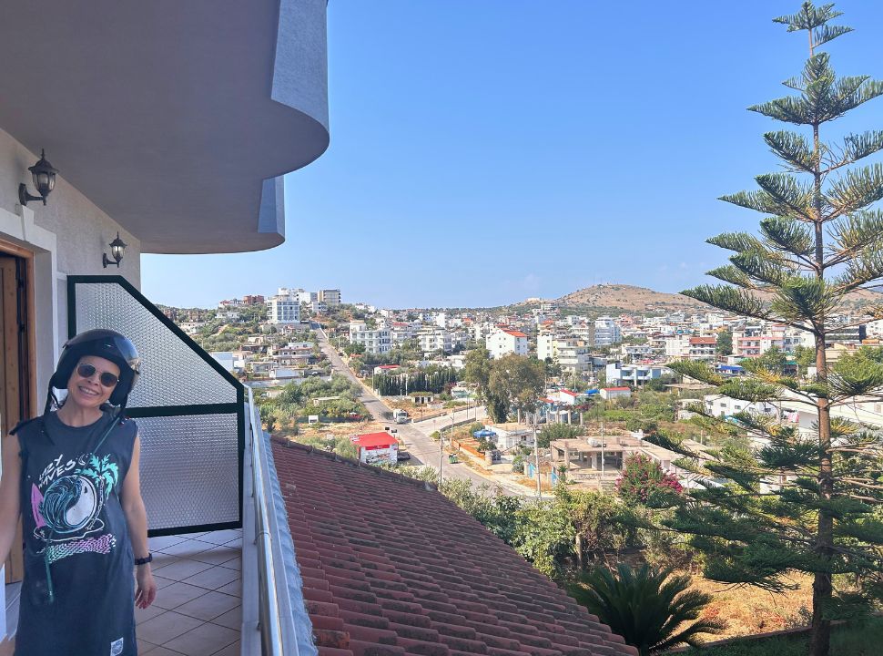 woman wearing a motor scooter helmet, standing on a balcony with view of the town Ksamil. You can see a lot of buildings