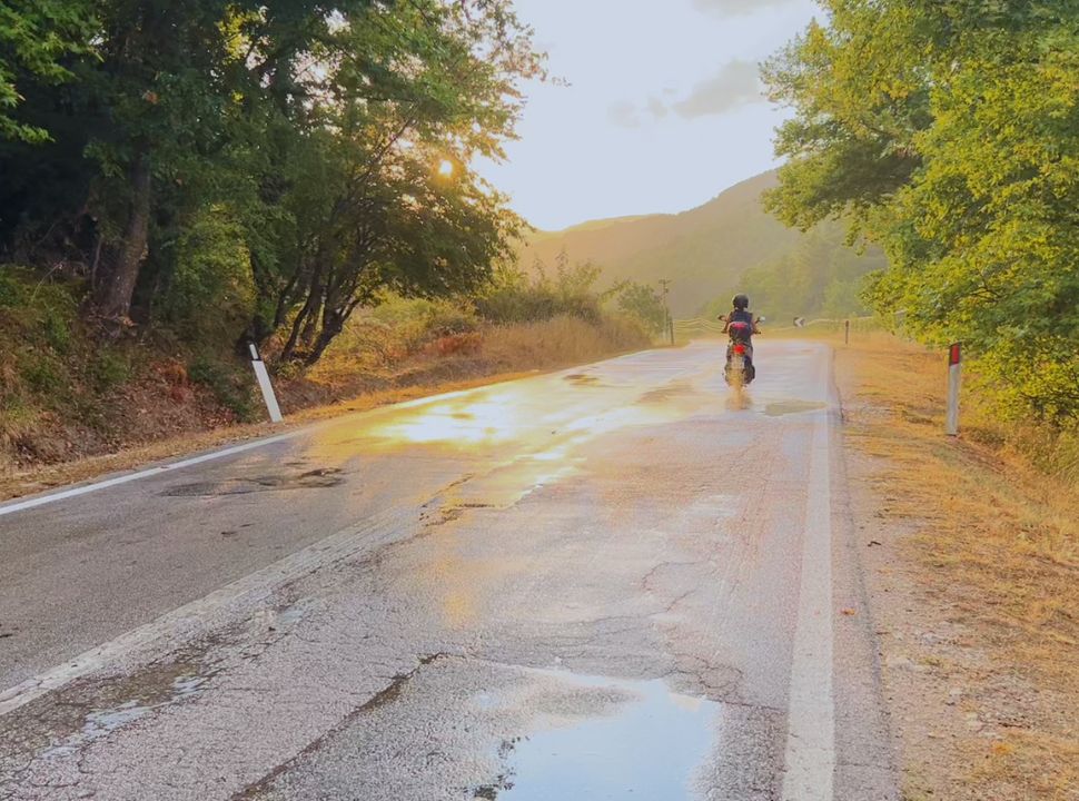 woman driving a scooter along a road where it had recently rained, the sun is setting giving a nice golden glow on the road and surrounding trees in Albania