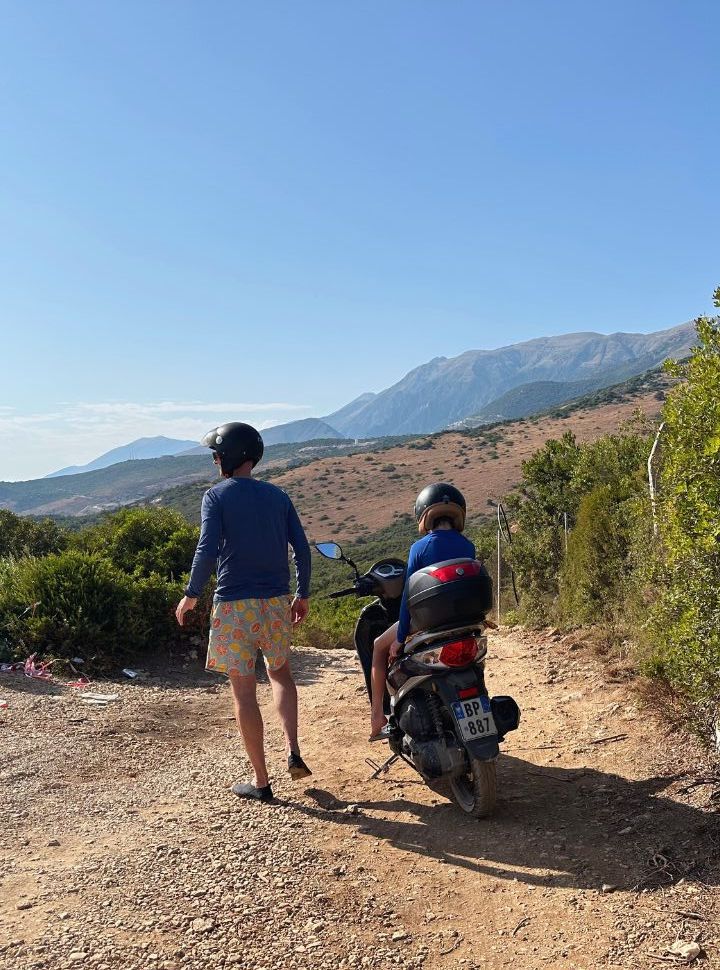 father and son taking a break of driving the motor scooter with mountain landscape in the back towards Aquarium Beach in Albania