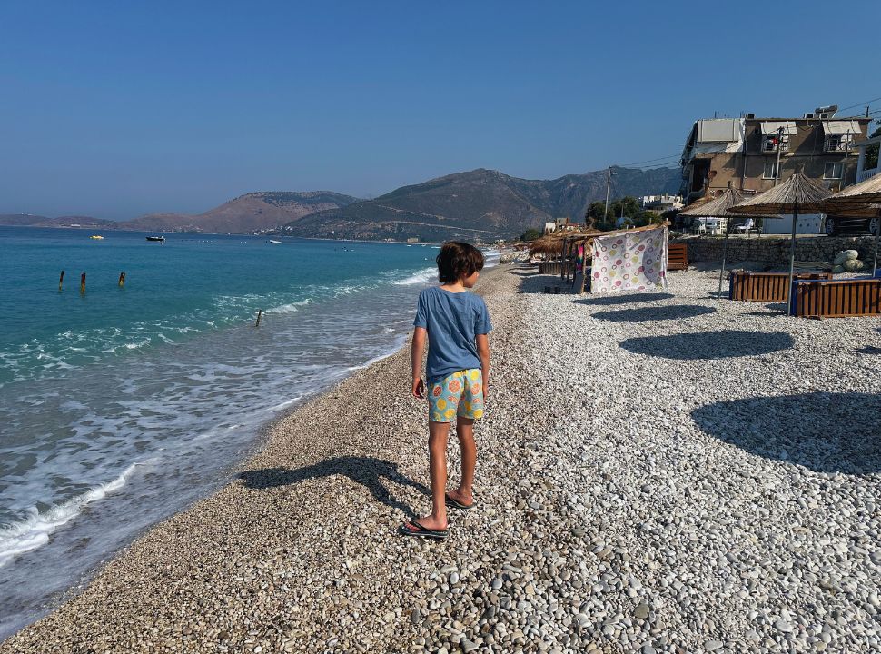 boy standing on the pebble beach with beautiful blue water, on the beach there are umbrellas that provide shade at Borsh beach Albania