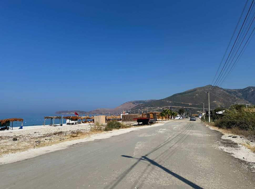 road along the beach with one side a couple of thatched roofed umbrellas, and on the other side bushes, with no buildings etc. at Borsh beach Albania