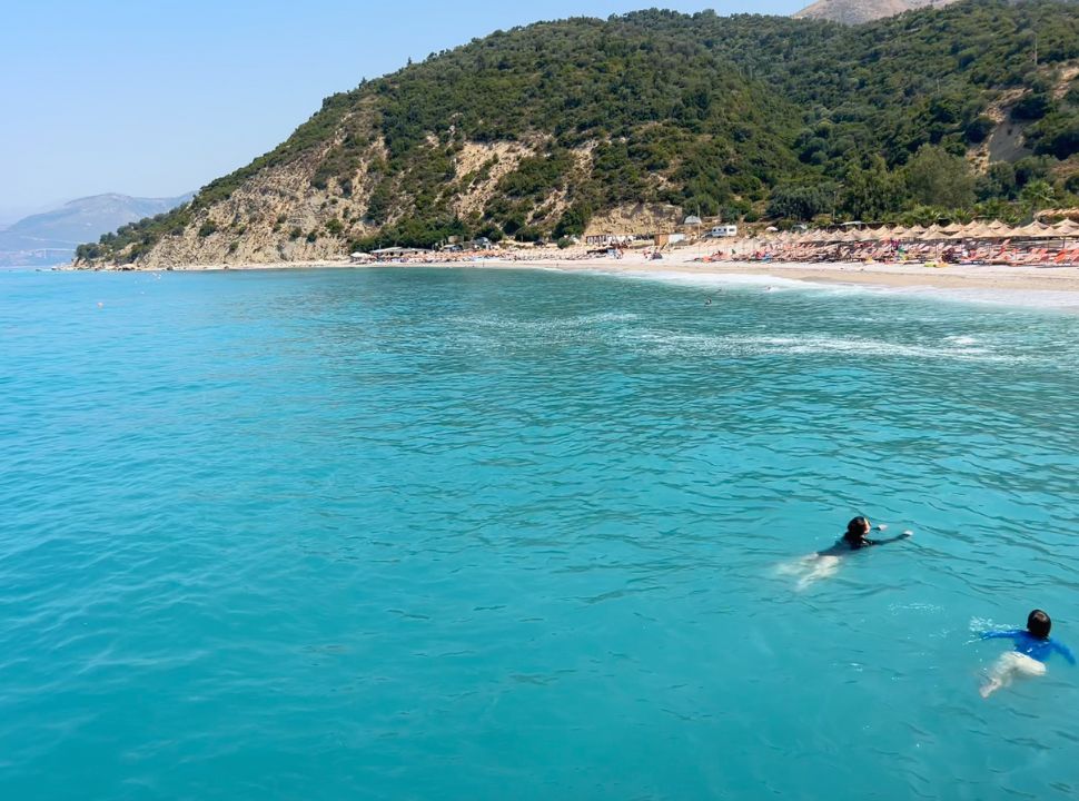 mother and son swimming in milky blue water, Buneci beach is set within a bay and has rows of sun beds 