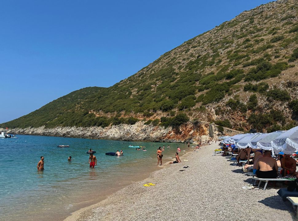 row of sun beds along the beach water front, with people swimming in the clear ocean water in Gjipe Beach Albania