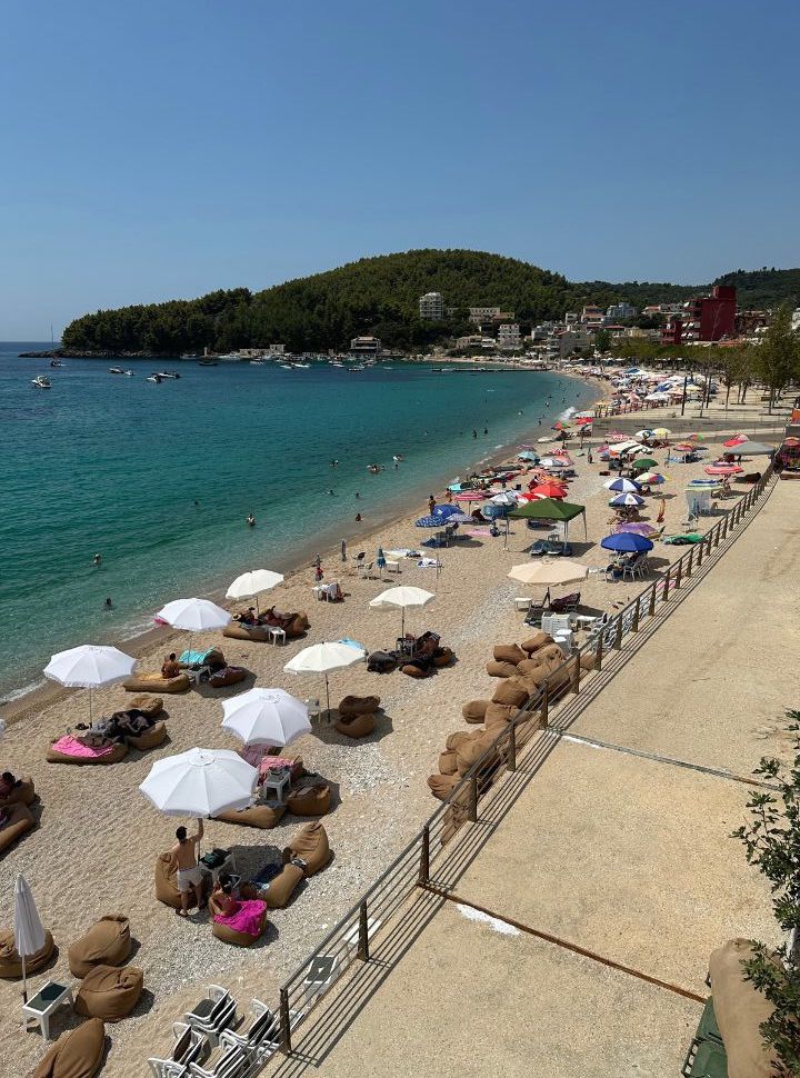 town beach with row of umbrellas and bean bags, people swimming in the clear turquoise blue water in Himare Albania