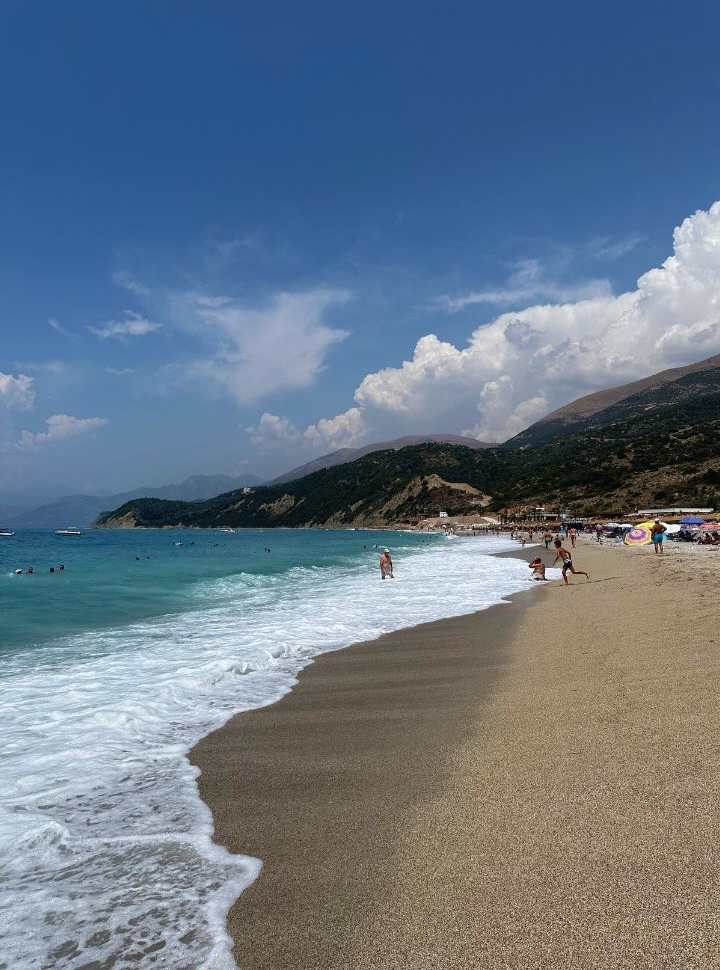 view of Lukove beach waterfront with people standing in the water, and in the distance umbrellas and the impressive mountains along the Albnaian Riviera. 