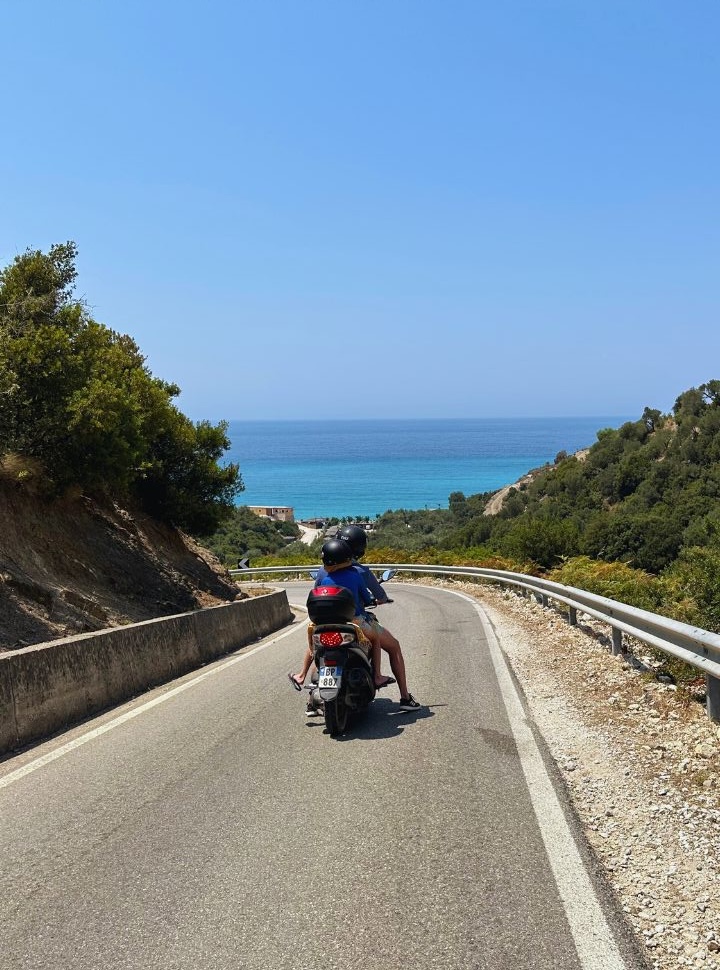 father and son on a motorscooter ready to drive down the road to the beach, ocean is visible in the distance at Lukove beach at the Albanian Riviera