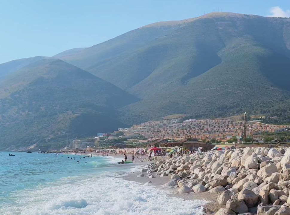 view of a beach with large white rocks, and in the far distance sun loungers, lots of accommodation and the impressive mountain slopes of the Albanian Riviera