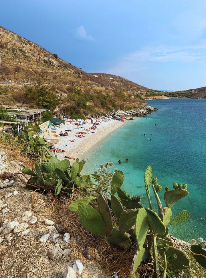 small patch of beach along the main road with a restaurant, people are lying on the beach and swimming in the turquoise water