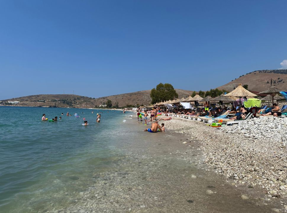 Qeparo beach where people are swimming in the water, others are sitting in the water shore or on the sunloungers under the umbrellas