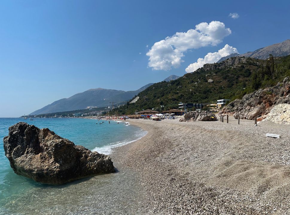 view of dhermi beach Albania, a long beach with pebbled sand and blue water, first section has no beach clubs, in the far distance there are sun loungers and restaurants