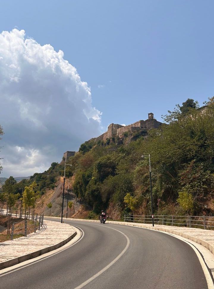 woman on a motorscooter riding a new paved road located around the old town of Gjirokaster, the castle is clearly visible. 