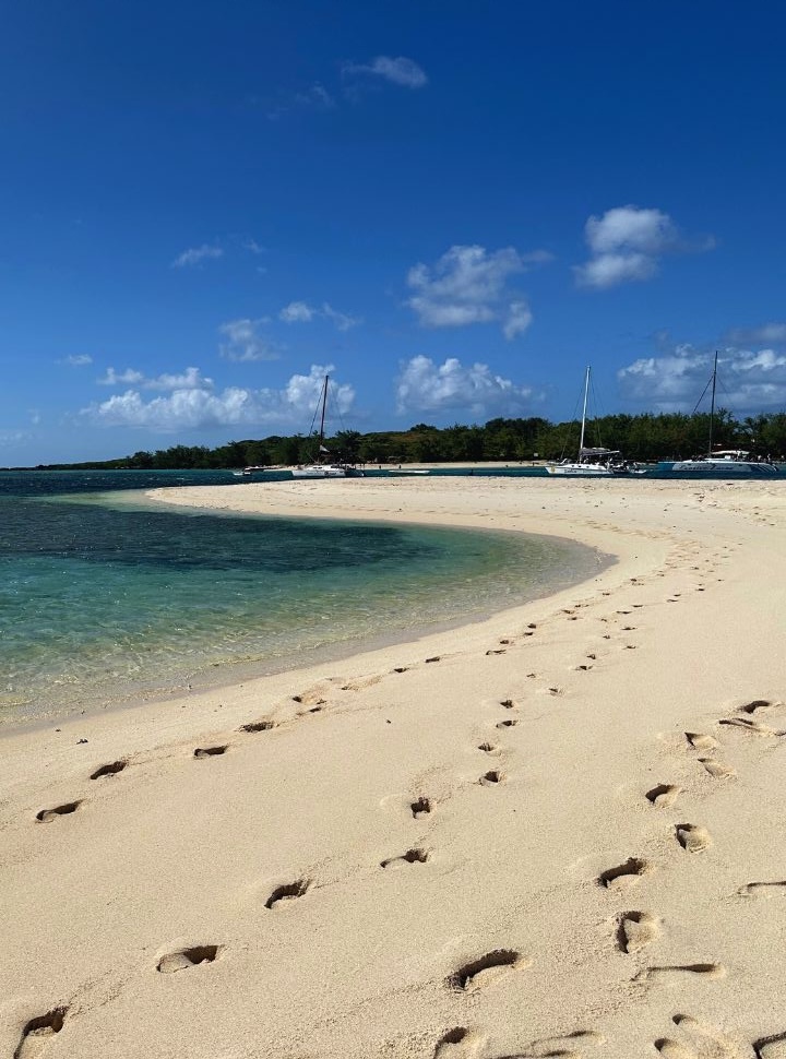 footprints in the soft sand of a tropical beach with emerald blue water with in the distance catamarans that take tourist out of a day