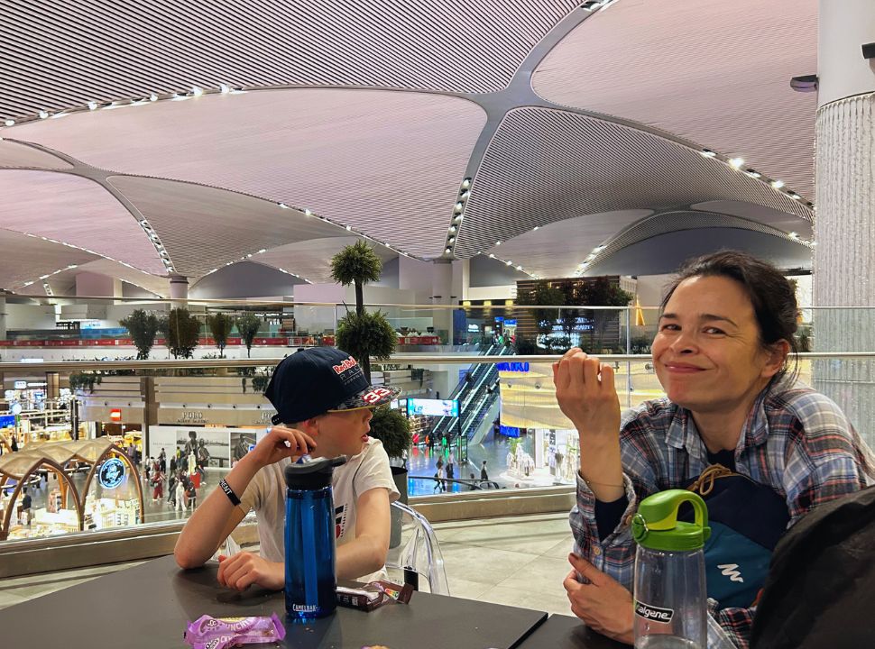 Mother and son sitting at a table at the Istanbul Airport, shops and waiting area are visible in the back.