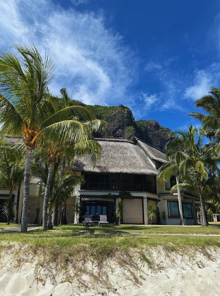 a room or bungalow at a beach resort with palm trees, thatched roof and le Morne mountain in the back
