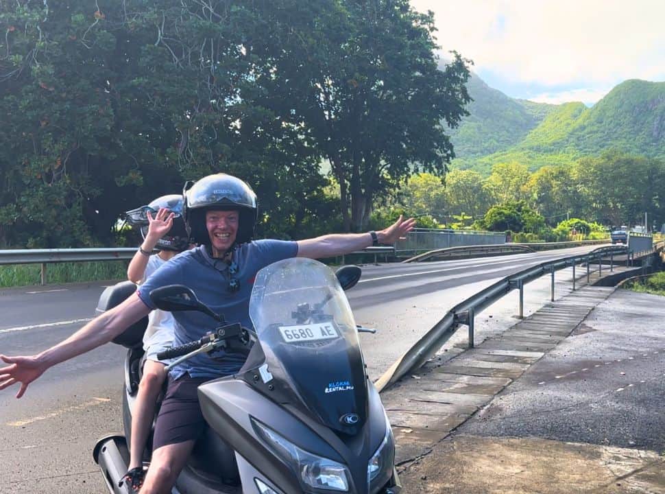 Father and son on a motor scooter standing on the side of a big main road posing for a picture, on the background is a mountain landscape with green slopes